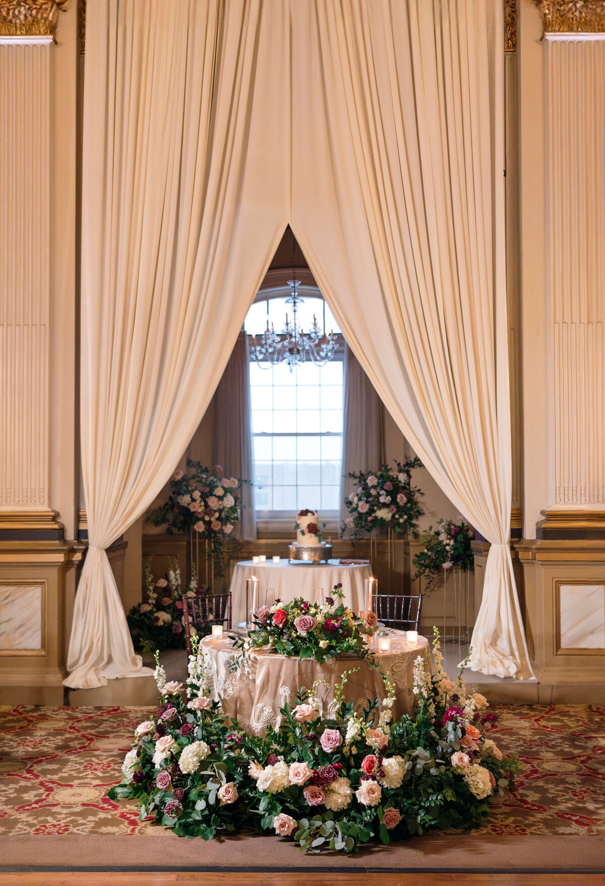 Elegantly decorated wedding venue with draped cream curtains framing a table setup. A small cake is on a round table in the background. Floral arrangements of pink, white, and green surround the foreground table, creating a romantic atmosphere.