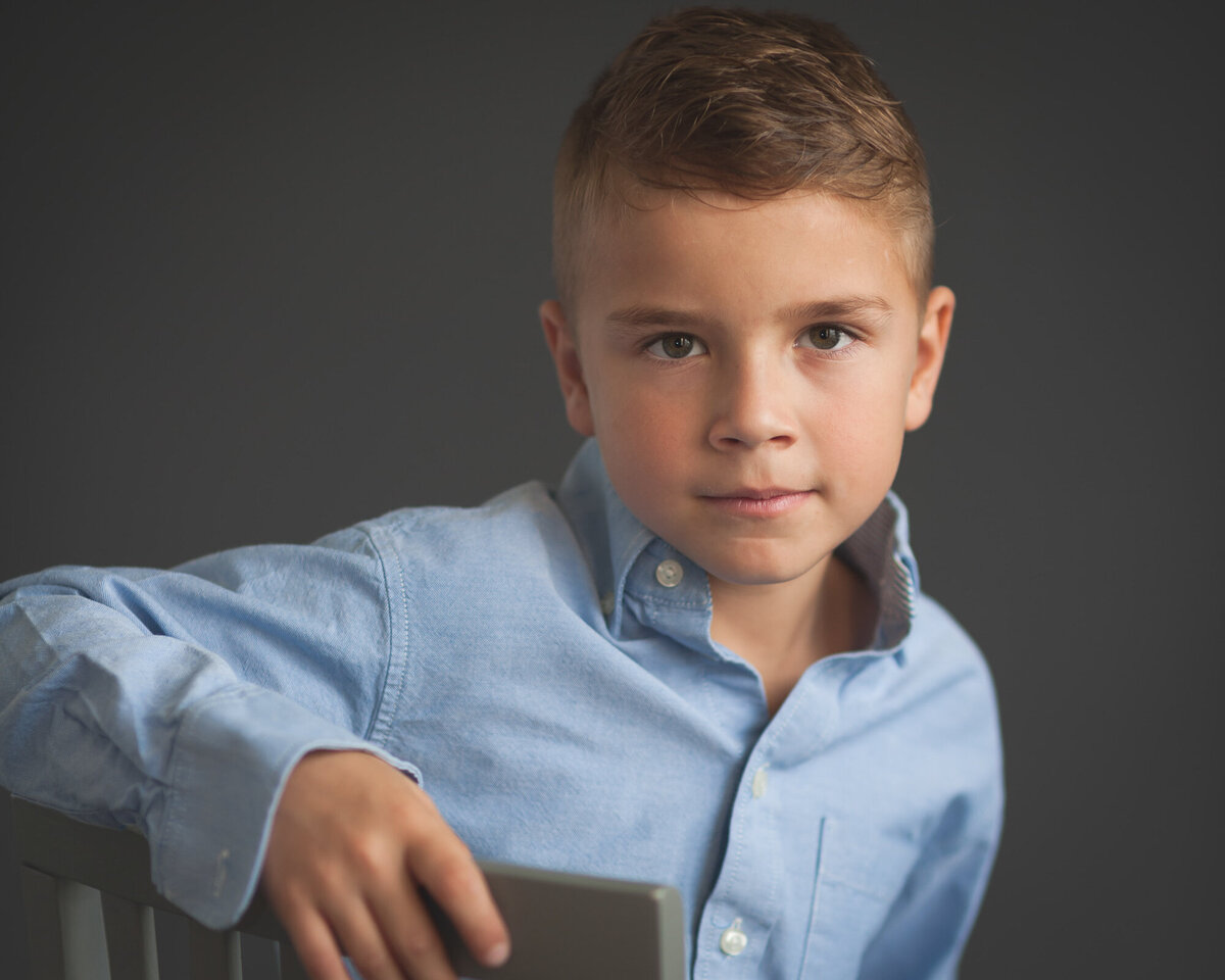 8 year old boy looking at camera with an intense gaze, grey background wearing blue dress shirt