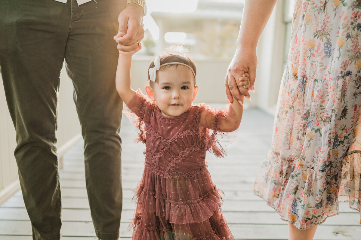 A girl walks towards the camera in golden hour light while holding her parents' hands.