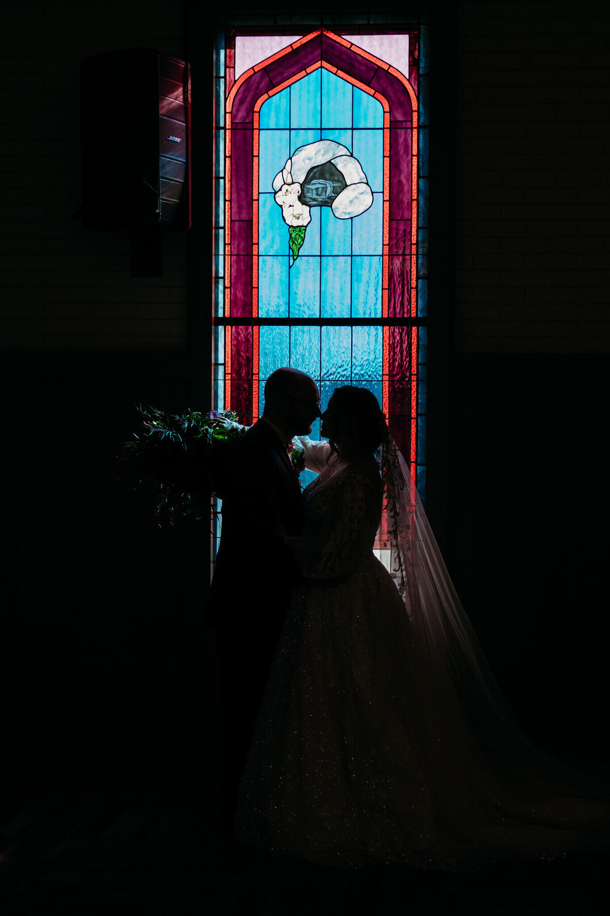 Photo of a bride and grooms silhouette standing in front of a stained glass window