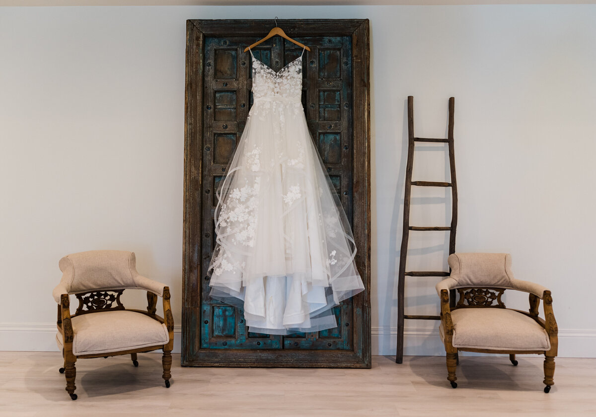 wedding gown with floral details and a soft tulle skirt hanging from a dark brown and blue door in a bridal suite surrounded by lounge chairs photographed by denver wedding photographer