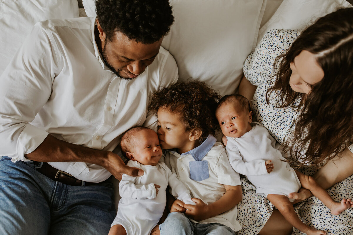 Mixed race bay area family with twin newborns snuggling on bed in home.  Sibling kisses newborn brother.