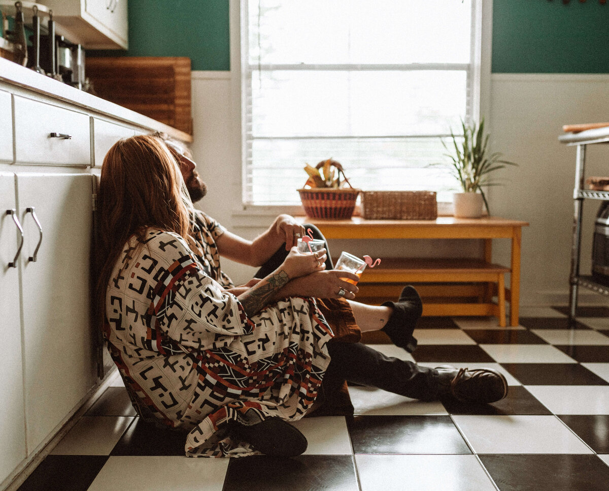 couple chats & drinks whiskey on retro kitchen floor during in - home engagement photo session