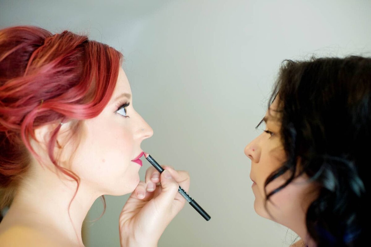 a makeup artist applies lipstick to a bride with bright red hair