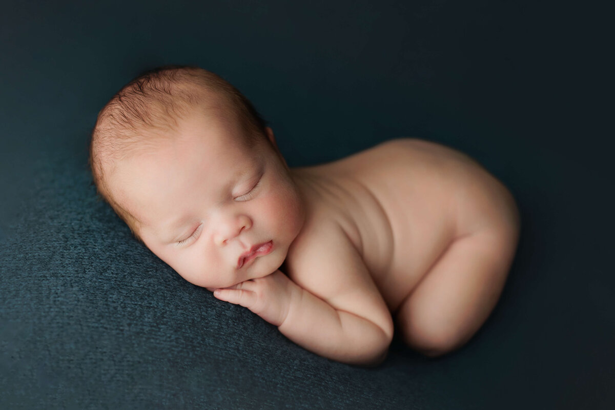Newborn baby laying on his tummy curled up n a teal backdrop at his newborn photography session at a dulles va photo studio