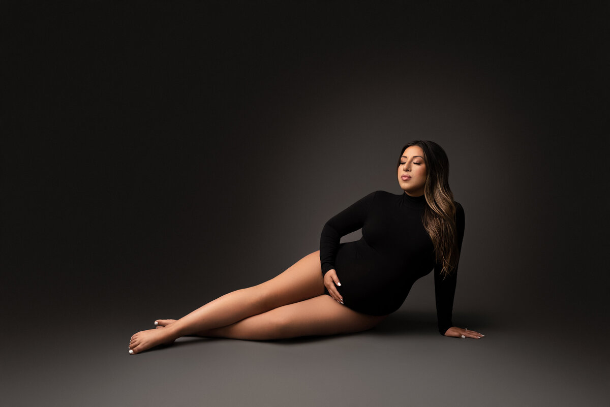 Maternity photo by Katie Marshall, New Jersey's premier maternity photographer. Woman in black long-sleeve bodysuit seated on the floor with legs extended and ankles crossed. One hand supports her weight, the other gently cradles her bump. She gazes towards the light source on the left.