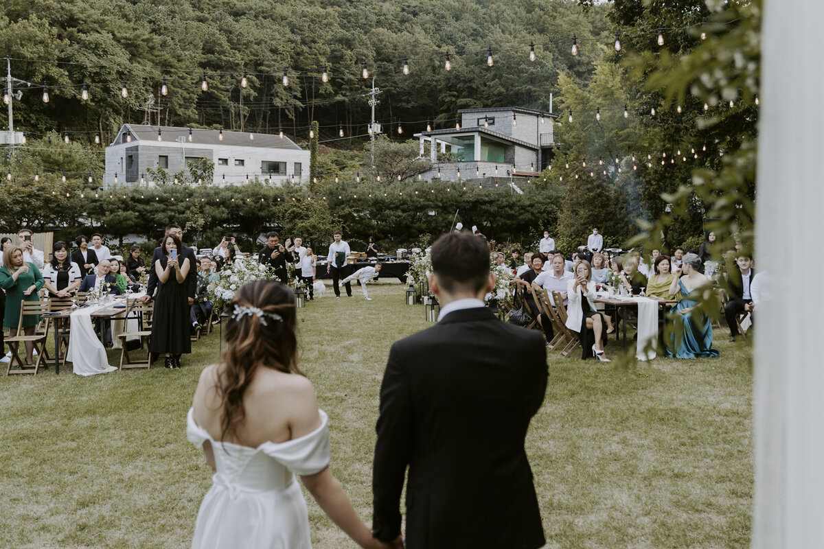 the couple hold hands in front of the guests in an outdoor ceremony in seoul