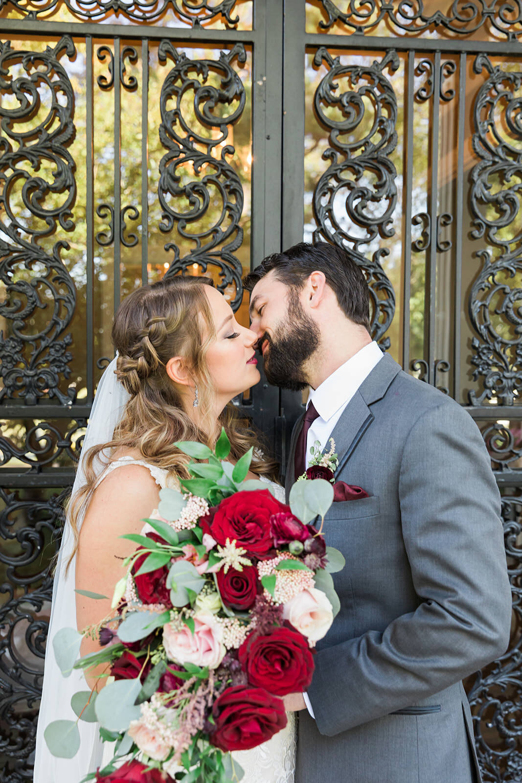 Bride and Groom kissing in front of iron door holding cascading bouquet with reds, blush, white, and greenery