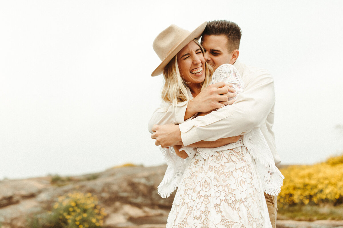 Groom wrapping his arms around the bride on top of a mountain with yellow flowers behind them
