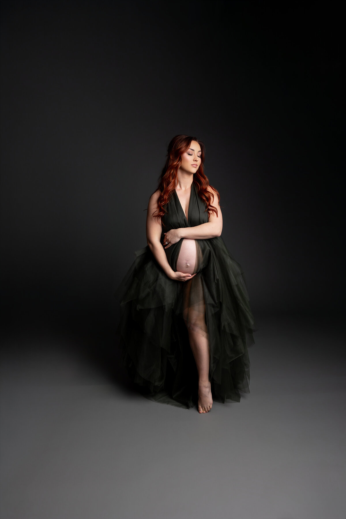 In a sleek black dress, she gently cradles her belly, her serene expression highlighted against a dark backdrop. Captured by Inland Empire photographer Kaitlyn Dawn Photography.