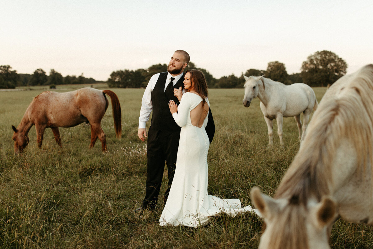 Couple in wedding attire standing in a field surrounded by horses