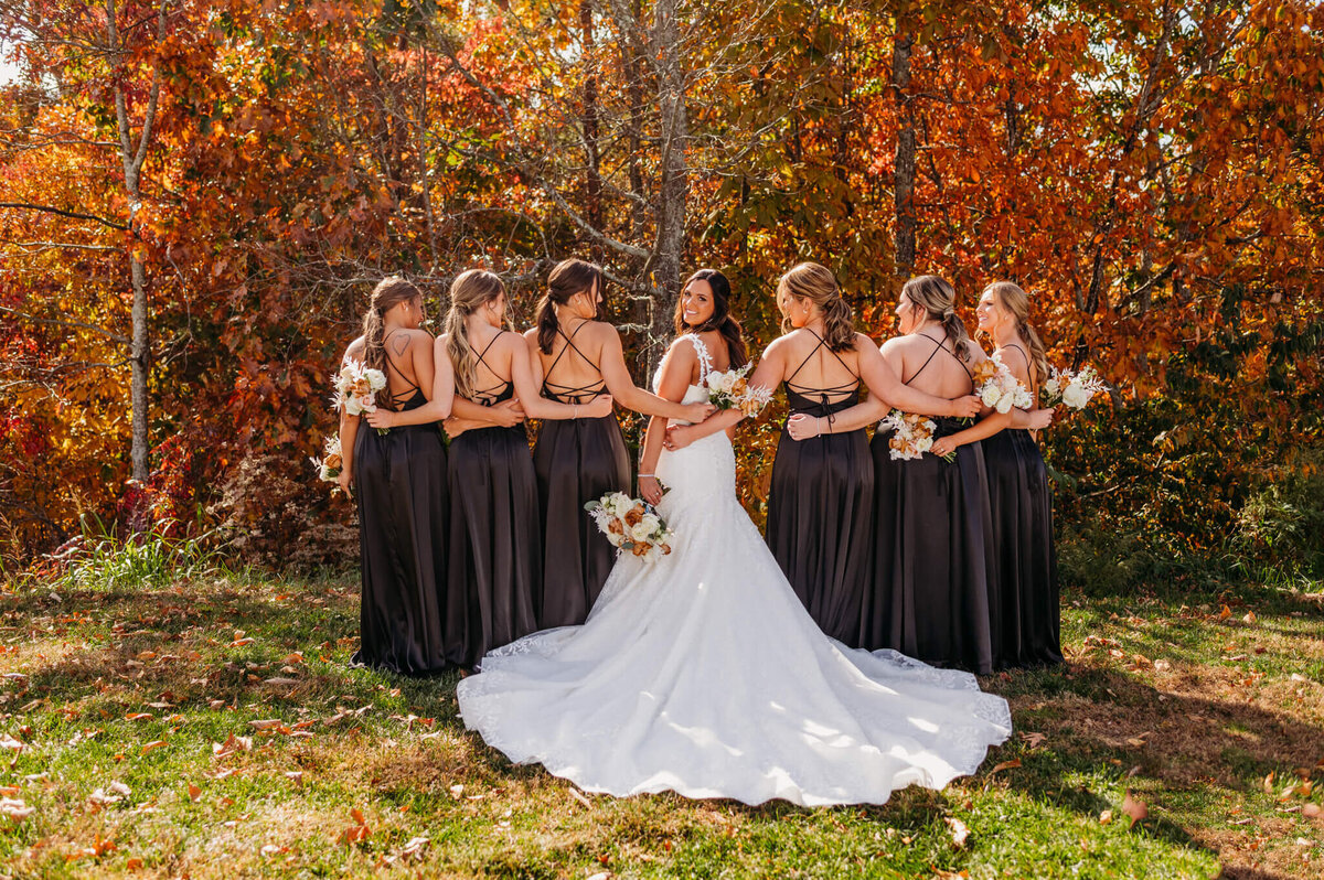 A photo of a bride and her bridesmaids wearing black standing in front of fall foliage