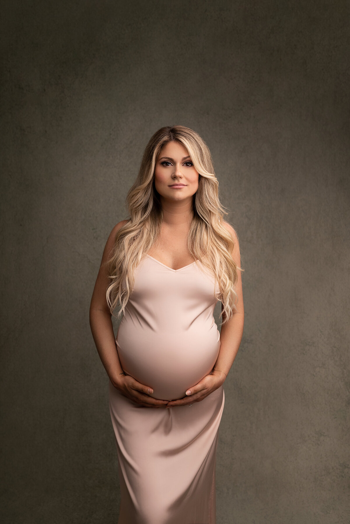 New Jersey's best maternity photographer Katie Marshall captures expectant mom for fine art maternity photos. Mom is standing in a long pink silk maternity slip facing the camera. Both of her hands are underneath her baby bump. She is looking at the camera with a closed-mouth smile.