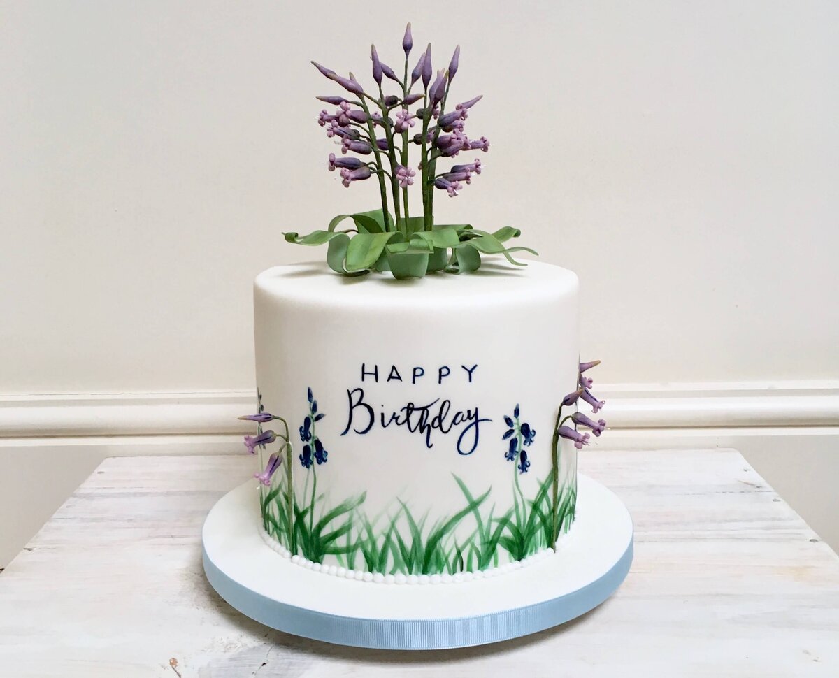 A birthday cake with hand painted flowers as well as sugar flowers coming out of the side and top of the cake