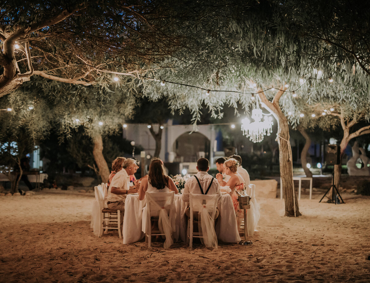 A small party sit around a table set on the beach under a dark sky decorated with festoon lighting