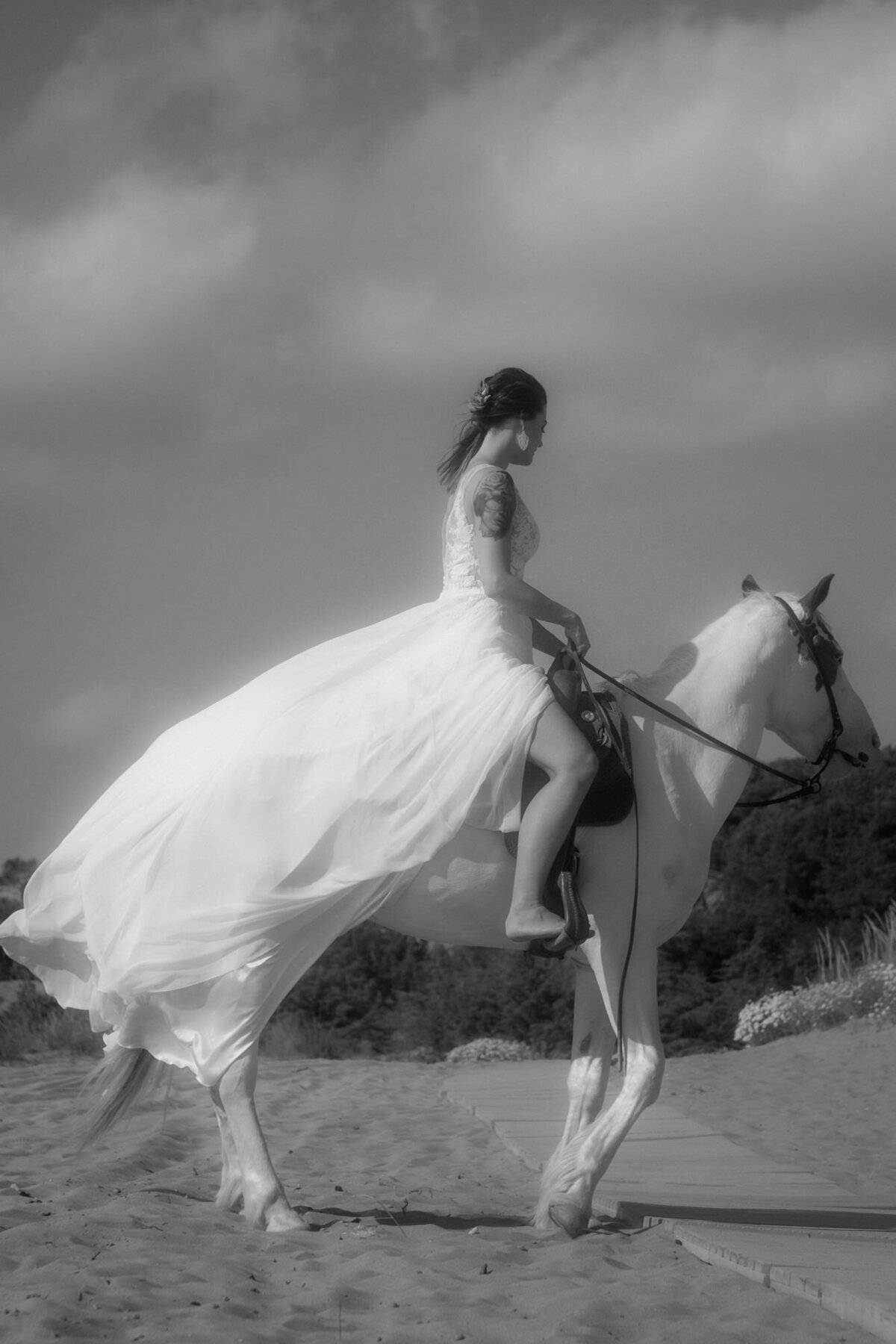 Von der Seite betrachtet sitzt die Braut barfuß auf einem Pferd am Strand.
