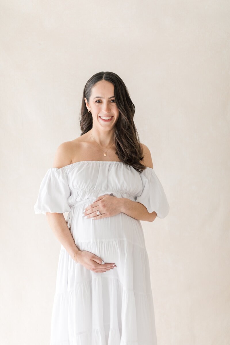 A pregnant woman wearing a white off the shoulder dress holds her stomach as she smiles at the camera.during her Larenceville NJ maternity photography session