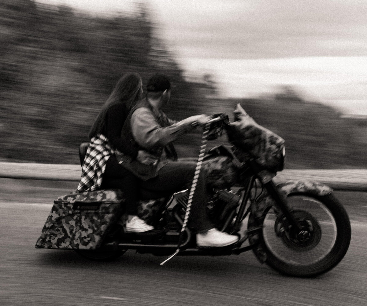 Girl with flannel tied around her waist sitting on the back of a motorcycle behind her husband while they drive down the street