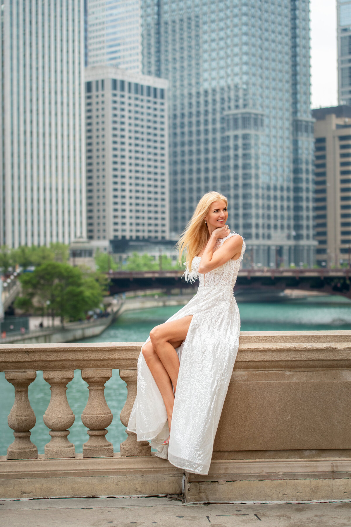 A wife to be is sitting on the rail by the Chicago river with skyscrapers in the background
