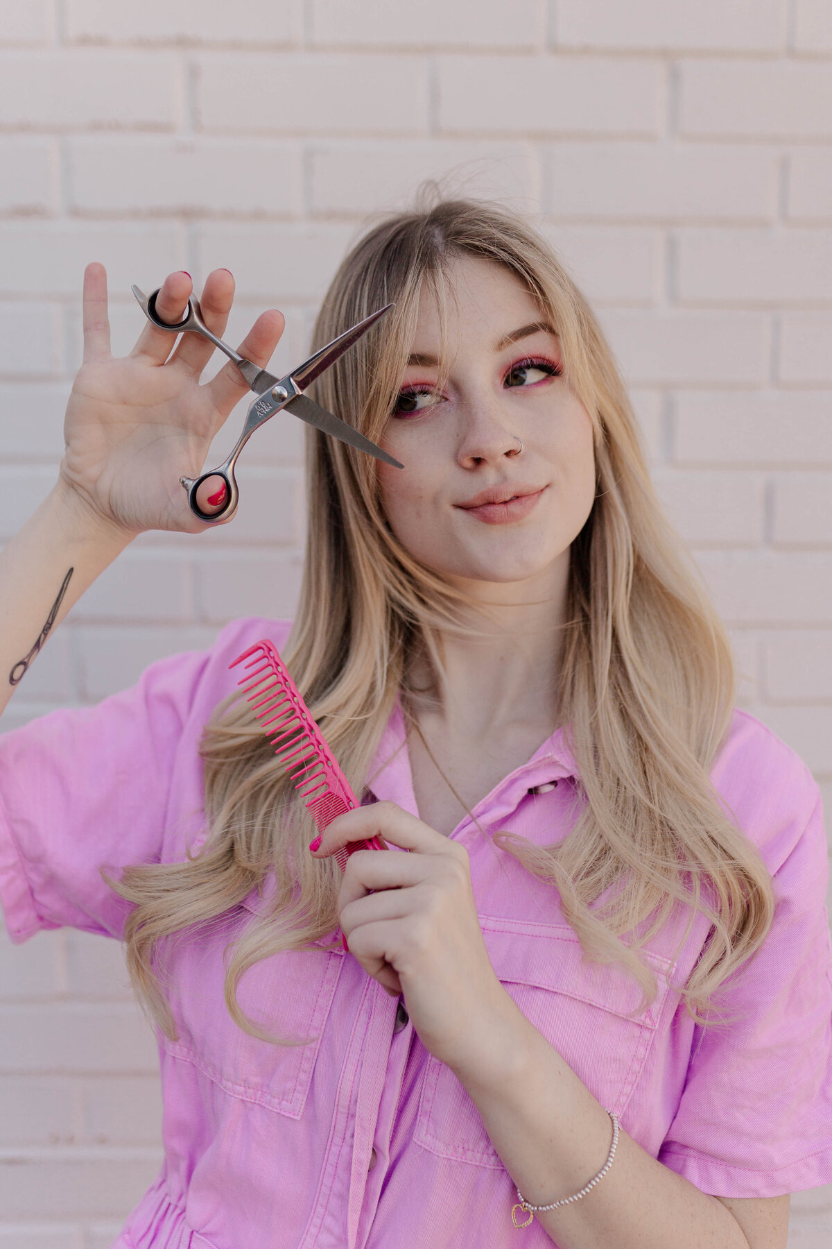 Branding photo for a hairdresser posing with shears and a comb wearing all pink in front of a white brick wall