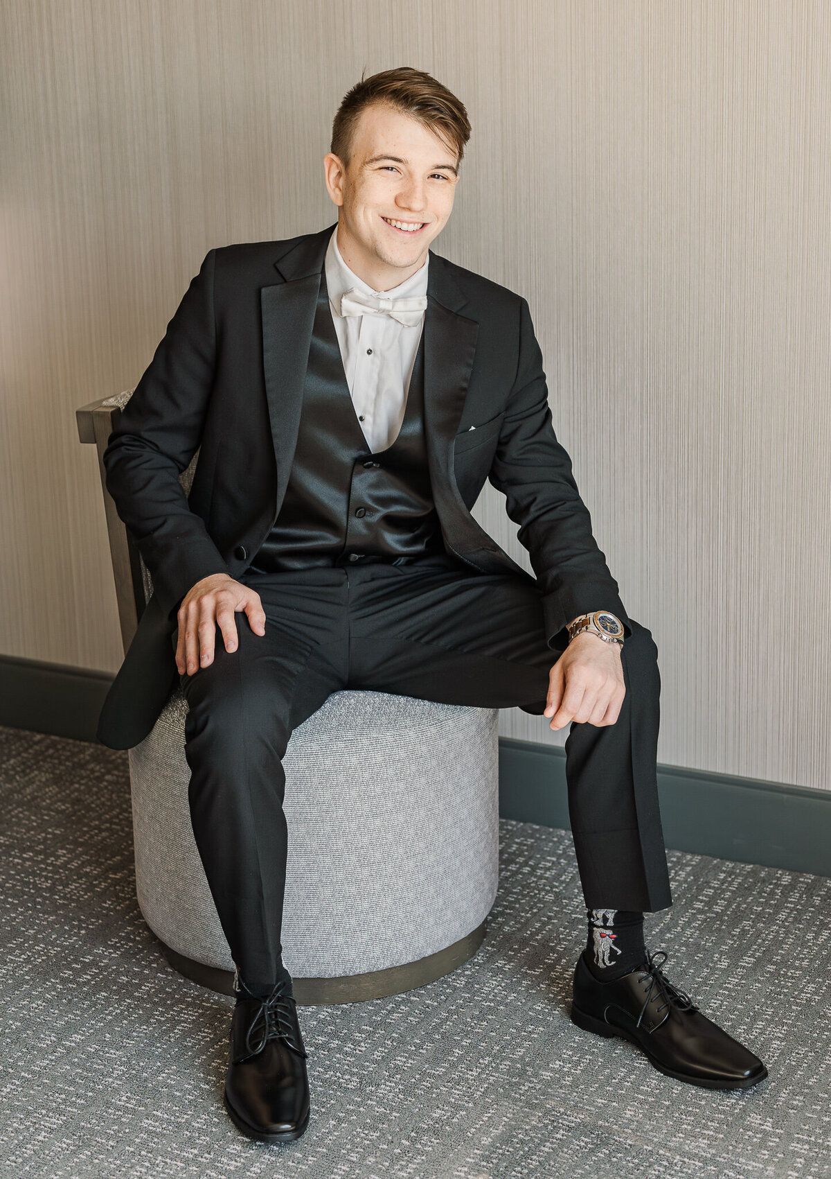 A groom sitting in a chair in his hotel before a wedding ceremony enjoying his Raleigh wedding photography session by JoLynn Photography