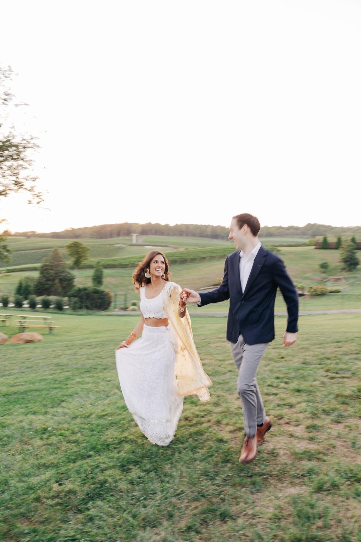 A joyful bride and groom holding hands and running across a grassy field with a serene landscape in the background.