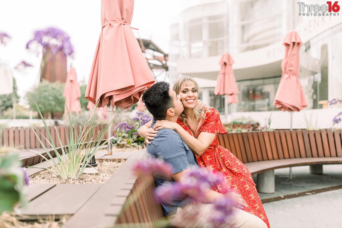 Bride to be is all smiles as her Groom kisses her cheek while resting on a bench