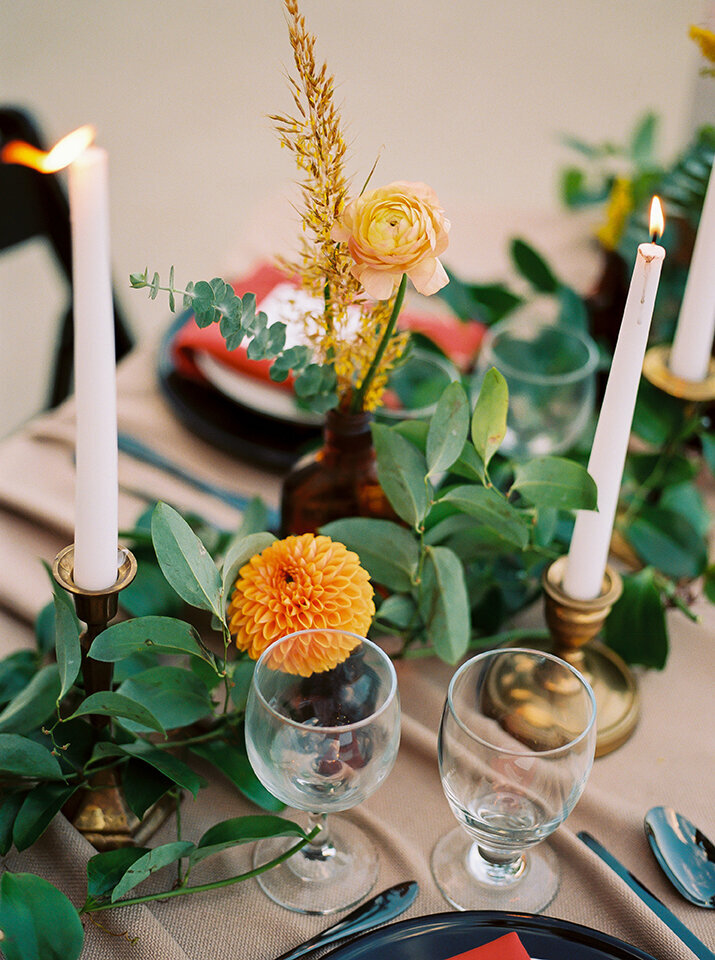 Table setting with green garland and candlesticks atop a tan table cloth with flowers and drinking glasses.