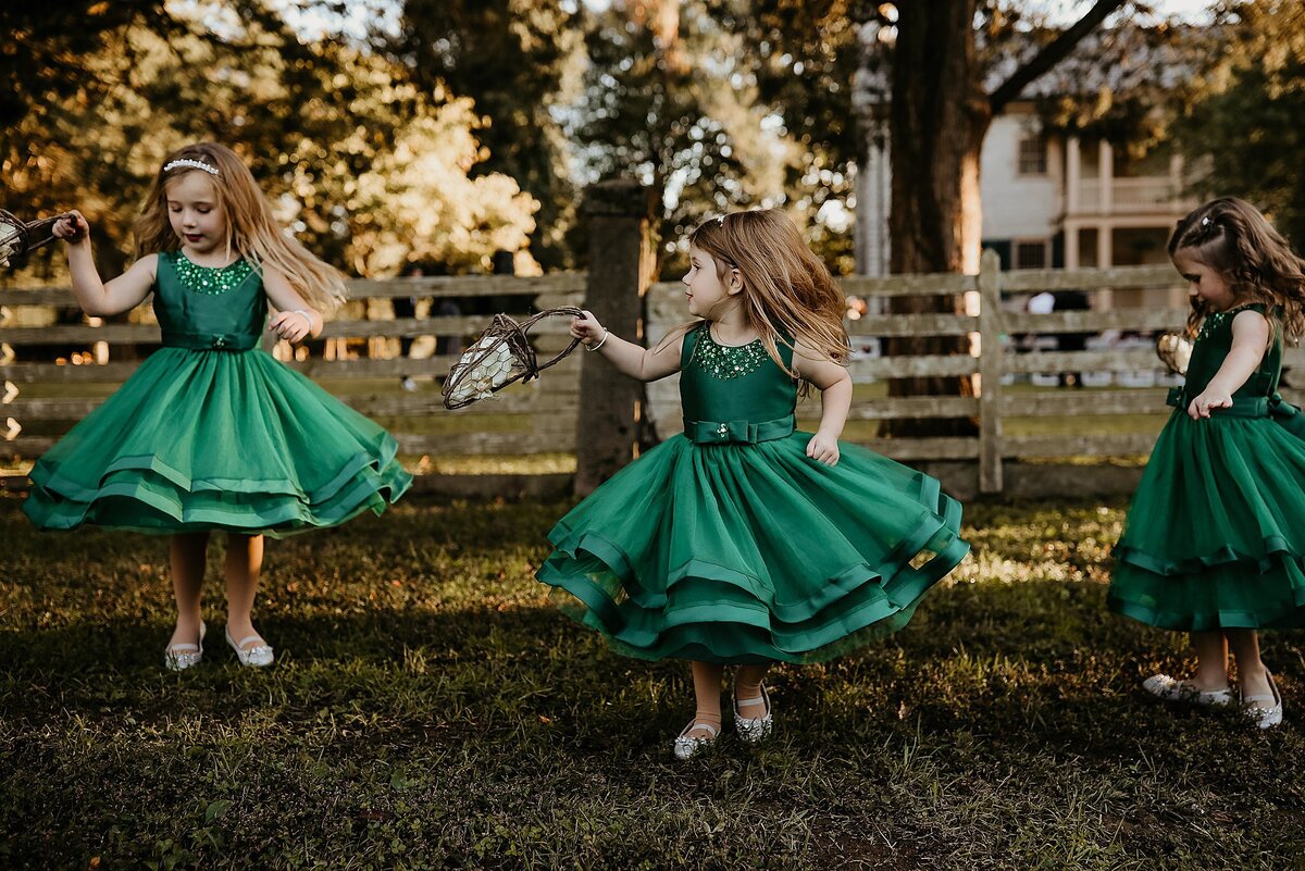 Three flower girls in emerald green dresses spin and twirl to make their skirts flare out as they dance down the aisle. The flower girls each hold a grapevine basket filled with white flower petals at Historic Rock Castle for a Nashville Wedding.