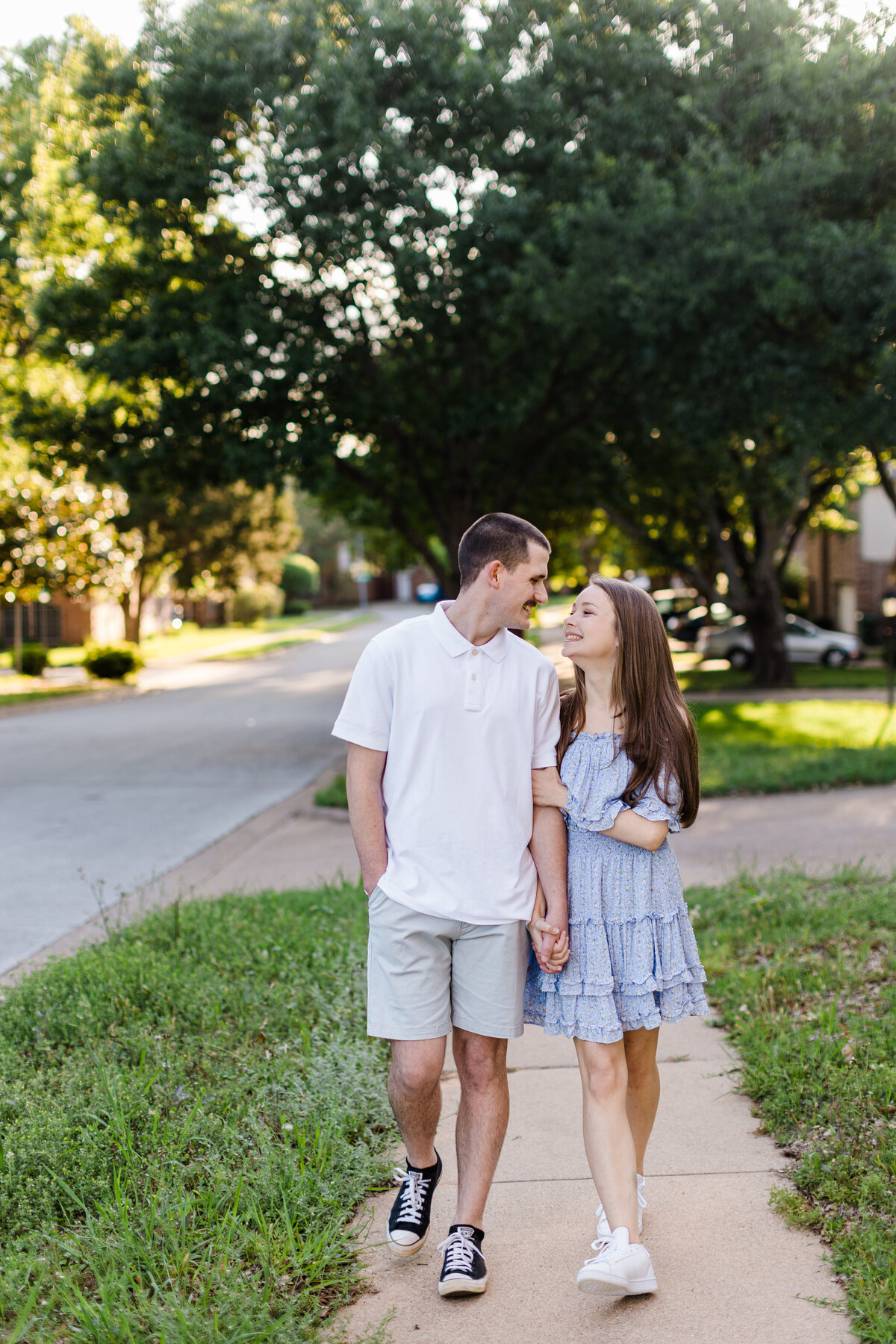 A couple holding hands and smiling at each other as they casually walk down the sidewalk during their engagement session in Grapevine, Texas. The woman on the right is wearing a short, blue dress while the man on the left is wearing a short sleeve, collared, white shirt and grey shorts.