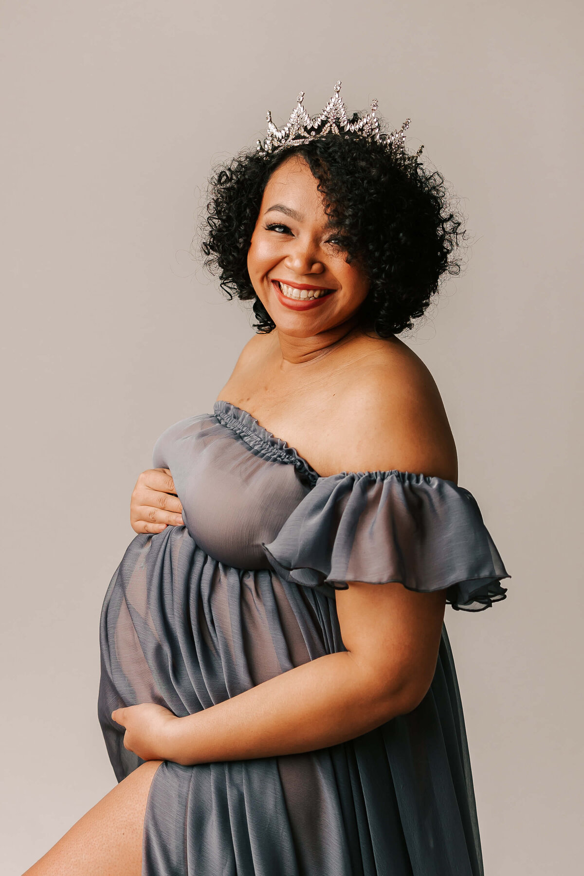 mom wearing blue dress and crown smiling for maternity portrait in portland studio