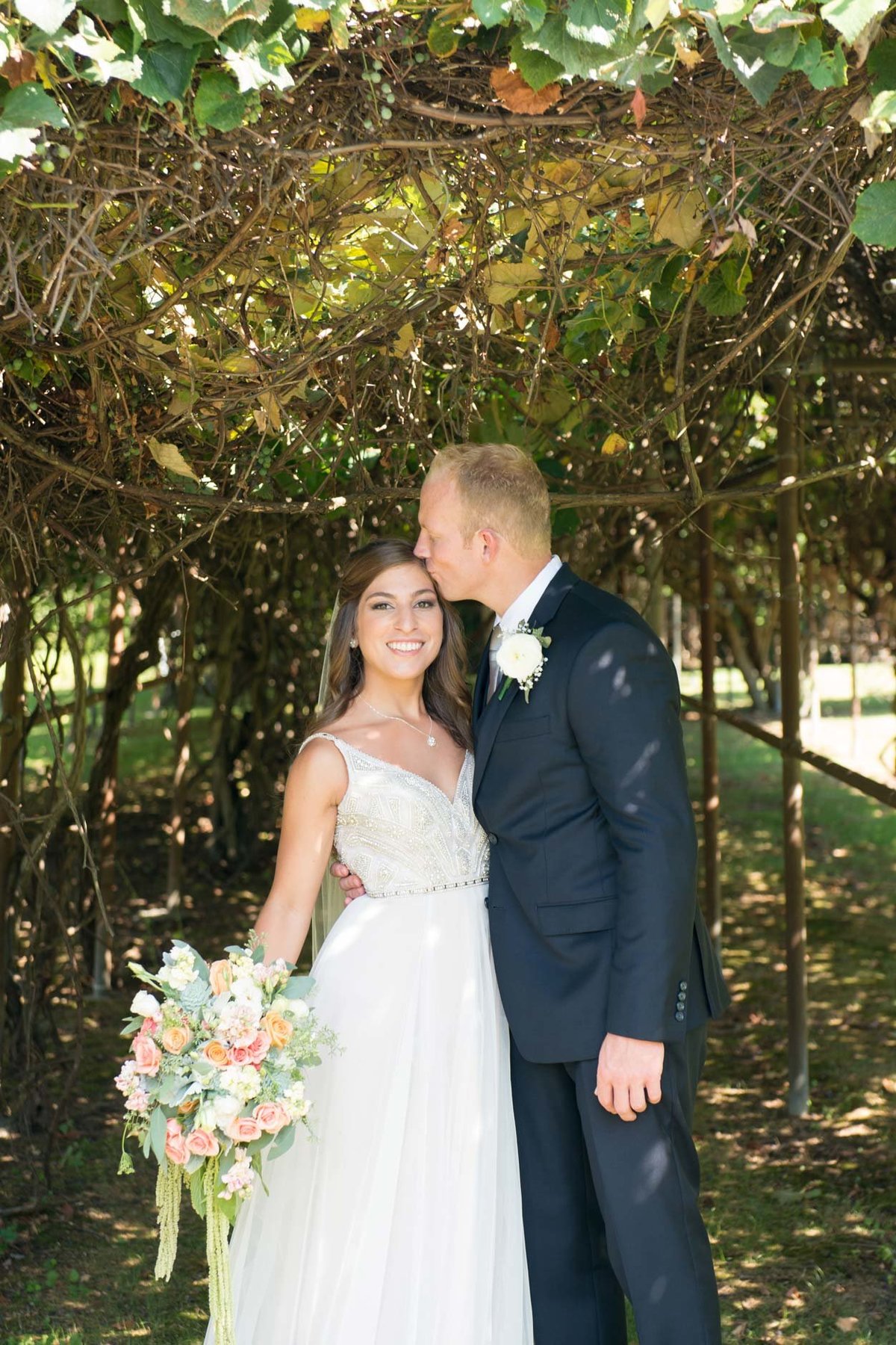 Bride and groom outside in trail under flowers with groom kissing bride at Flowerfield