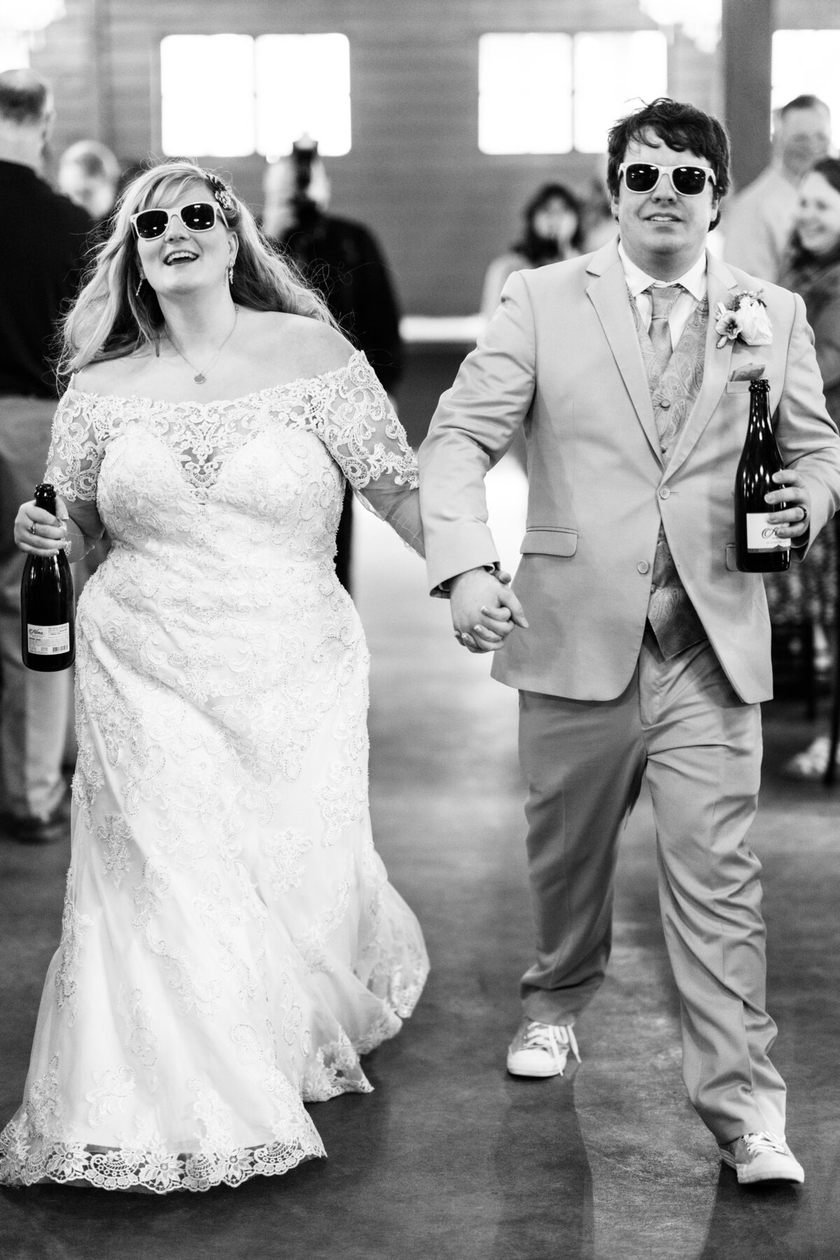 Bride and groom walk into wedding reception in sunglasses holding champagne bottles.