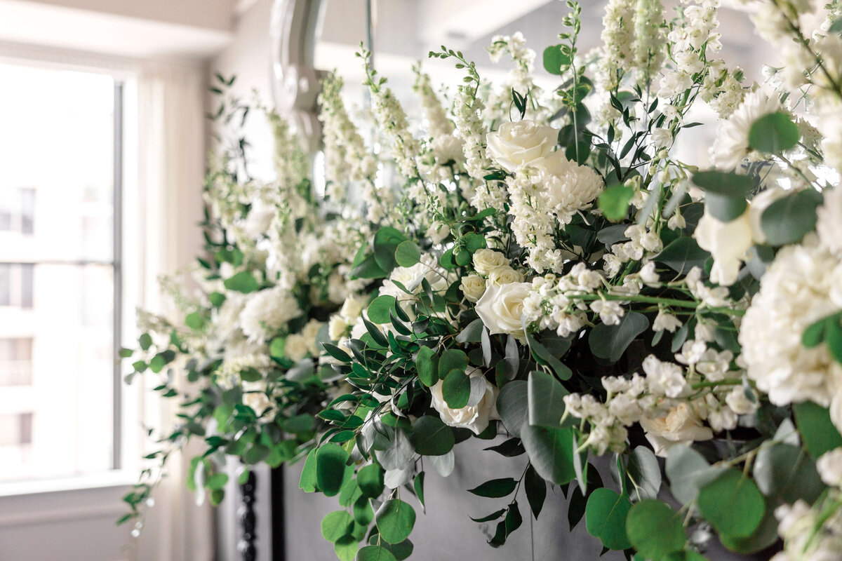 A lush arrangement of white flowers, including roses and delphiniums, is displayed against a bright window. The greenery includes eucalyptus leaves, creating a fresh and elegant contrast.