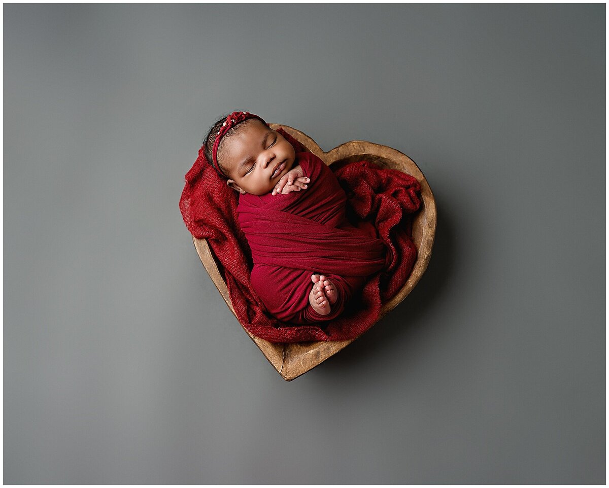 A newborn baby laying on a miniature prop heart in red wrap.