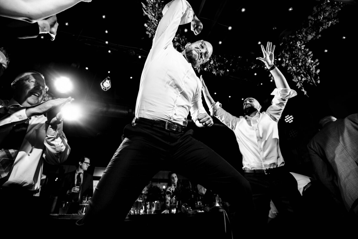Black-and-white-photograph-from-a-low-angle-perspective-of-a-groom-jumping-in-the-air-while-dancing-at-his-reception-at-Eventide-Brewing