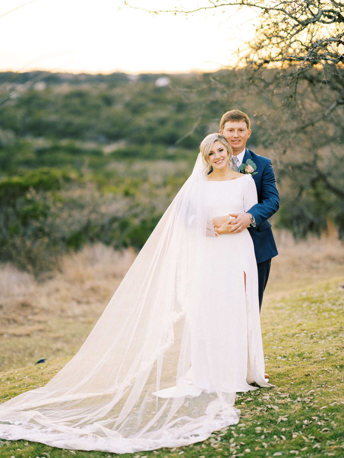 Bride with long cathedral veil is held by her husband in a meadow in Texas