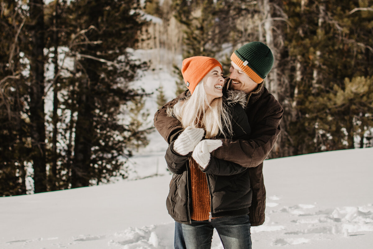 jackson hole photographers captures engagement pictures with forest behind an engaged couple as the man holds the woman shoulders as they stand in the snow for their winter engagement session