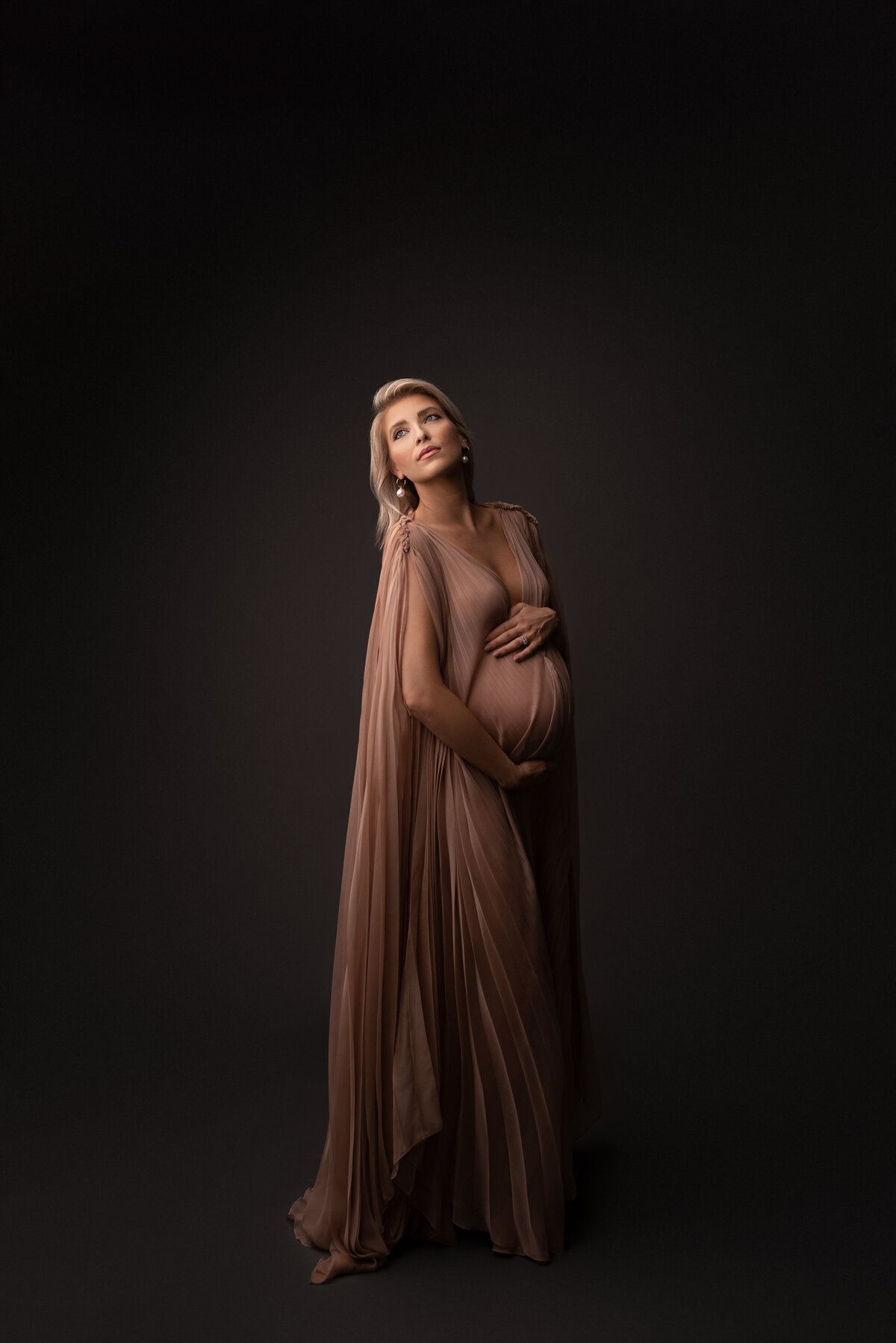Woman poses for fine art maternity photos with New Jersey's best maternity photographer Katie Marshall. Woman in long dusty rose maternity photoshoot gown with caped sleeves is standing angled to the camera. One hand is resting underneath her baby bump, the other overtop. She is looking over her shoulder to the light so her face is highlighted. Her body and bump are shadowed, creating dimension in the photo.
