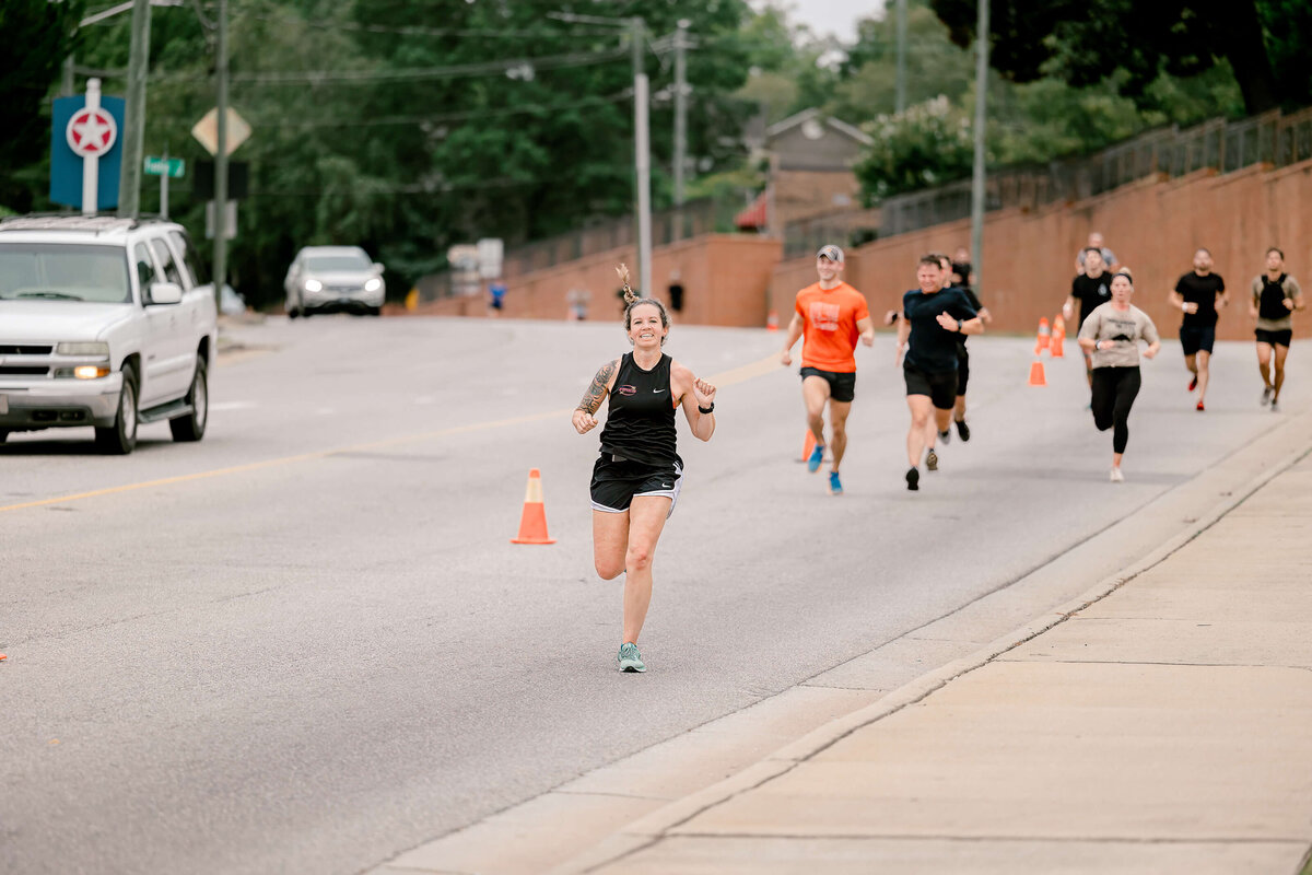 Several people are jogging on a road. A woman in a black tank top is in the foreground, smiling for an event photographer. Orange cones separate the runners from traffic in Fayetteville, NC. Cars are visible in the background, and trees line the road.