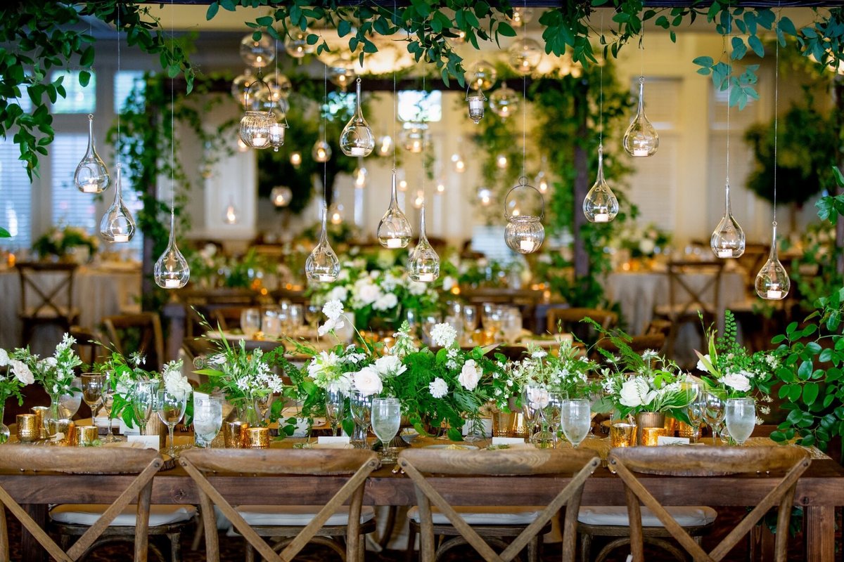 long wooden tables decorated with lush greenery centerpieces and hanging glass candles suspended from wooden trellis