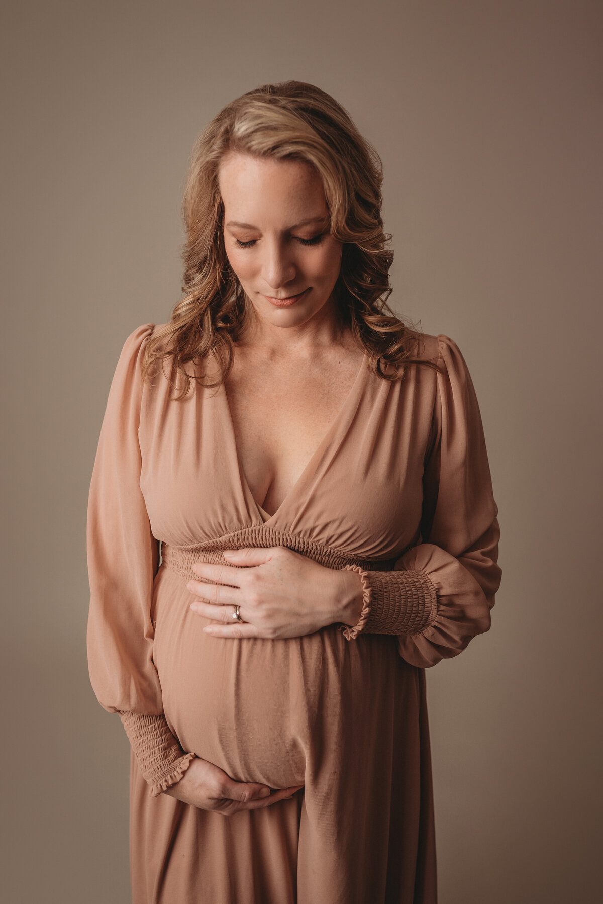 maternity photography marietta ga photo shoot for pregnant woman wearing light blush color dress holding baby bump with both hands looking at tummy posing on cream backdrop