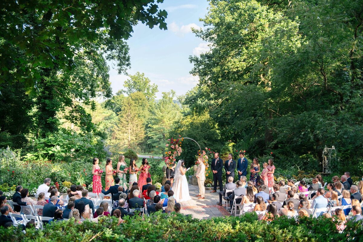 A wedding ceremony takes place outdoors under bright skies, surrounded by lush greenery. The couple stands at an arch adorned with flowers, with the bridal party in colorful attire. Guests are seated, witnessing the event amidst verdant surroundings.