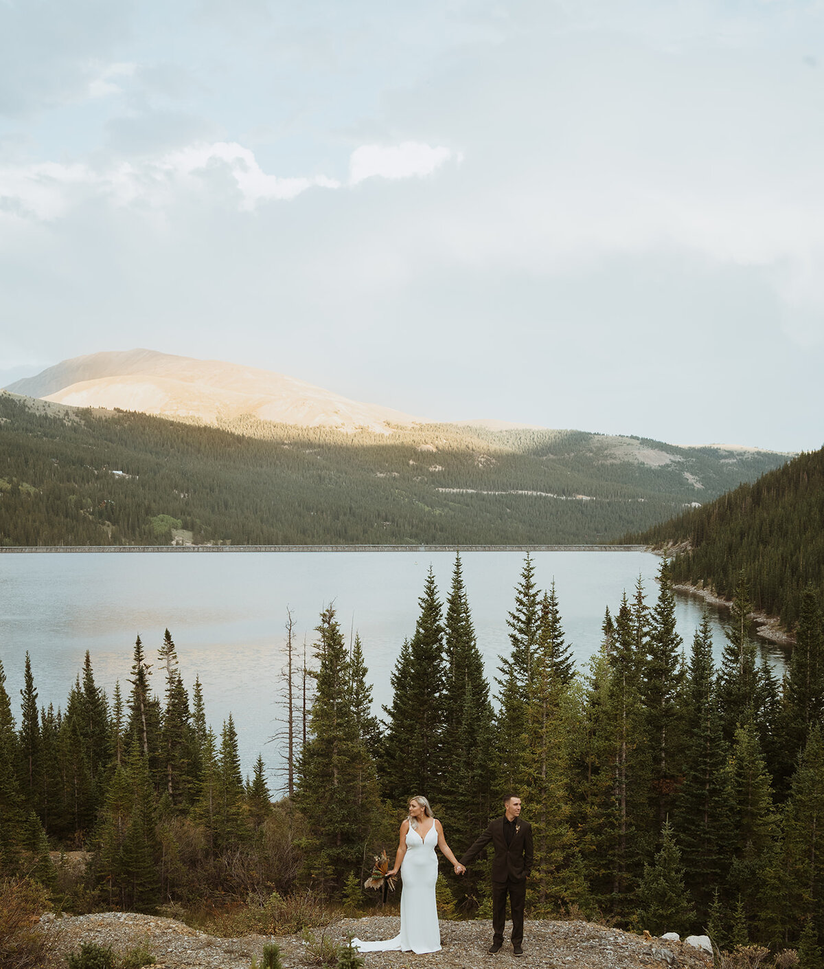 bride and groom are standing on the edge of the cliff kissing while the bride has her bouquet in the air with a large lake and pine trees in the background.