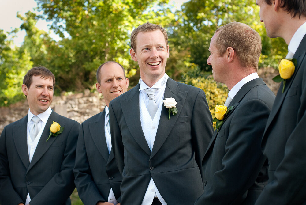 Red haired groom wearing a light grey cravat and dark grey tailcoat with white buttonhole laughing with his groomsmen