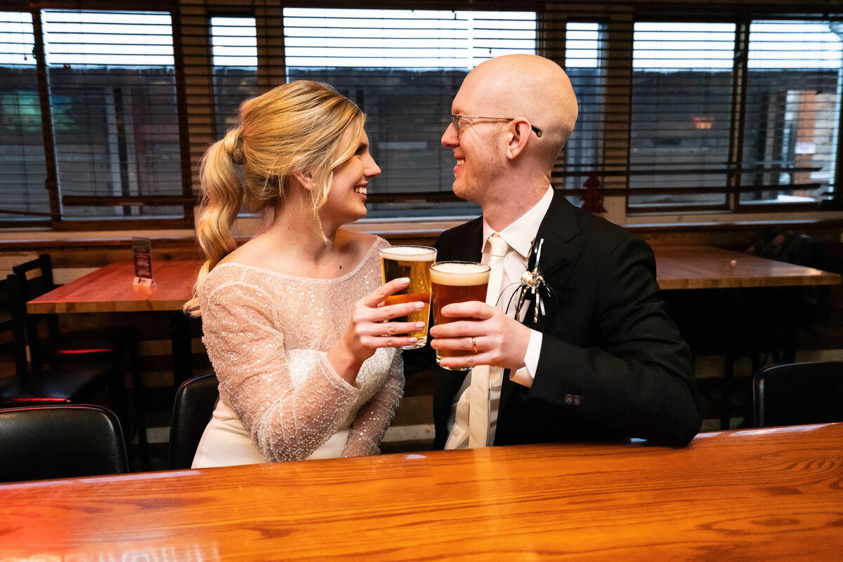 Bride and groom clink beer glasses at bar after wedding.