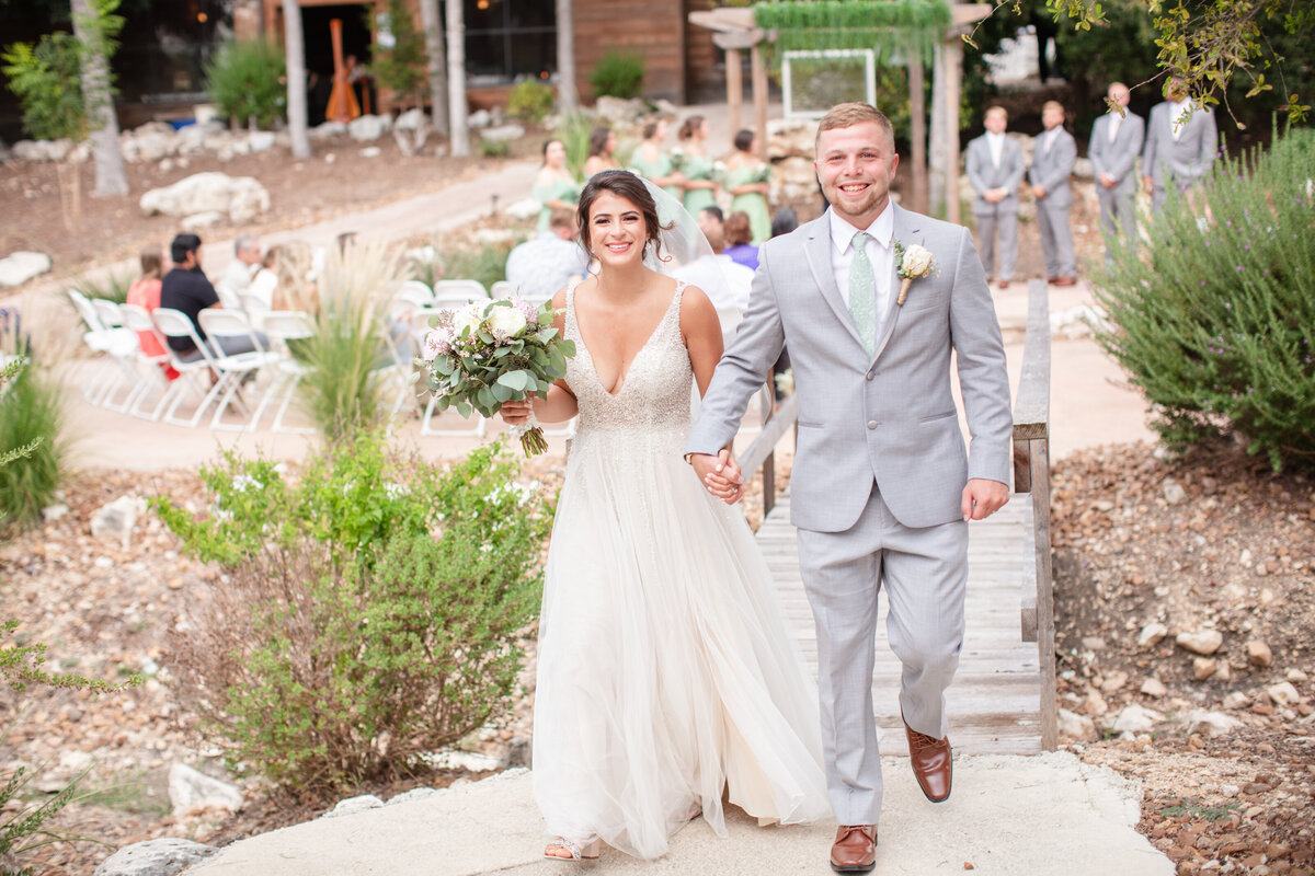 bride in v neck and groom in gray suit walk energetically from ceremony at Geronimo Oaks Texas wedding