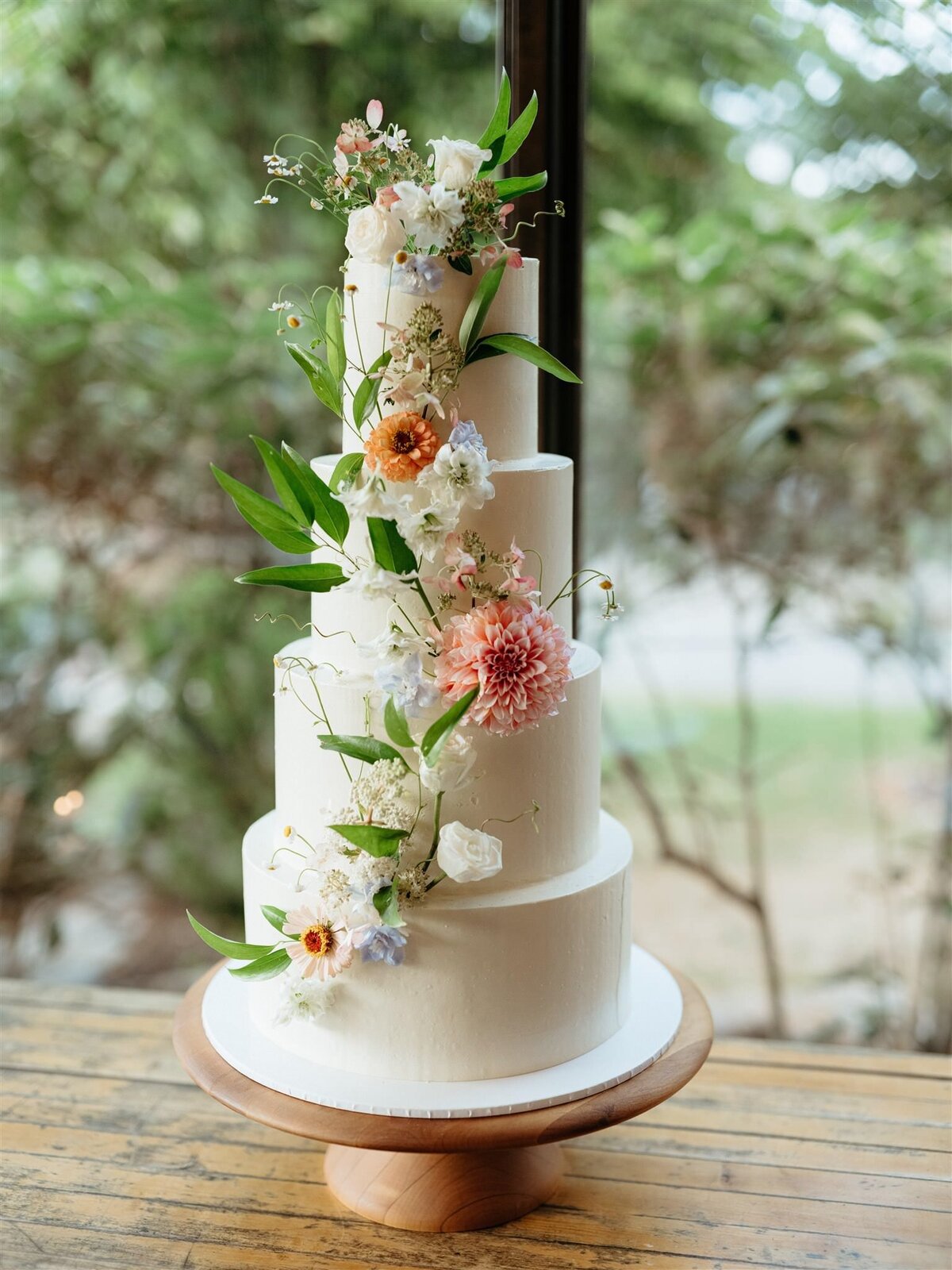 Romantic four-tier white wedding cake with whimsical and colorful floral accent sites on top of a simple natural wood round pedestal cake stand in front of large window overlooking trees and lake.