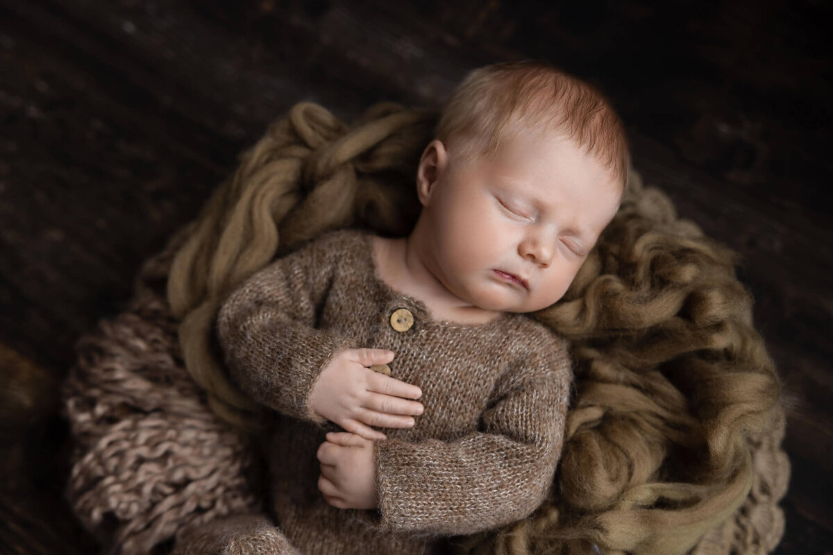 close up of newborn baby sleeping in a bowl wearing a brown knitted outfit