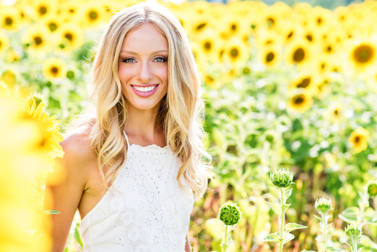 Blonde high school senior girl poses in sunflower field for her senior portraits.